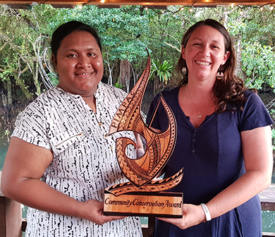 Two people holding a wooden sculpture of a traditional Polynesian fish hook smile at the camera. The base of the sculpture has the words Community Conservation Award engraved in it.